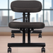 Boss ergonomic kneeling chair with black upholstered cushions and a metal frame, featuring adjustable height settings and caster wheels for mobility, positioned in an office setting with a view of a city skyline through the window in the background.